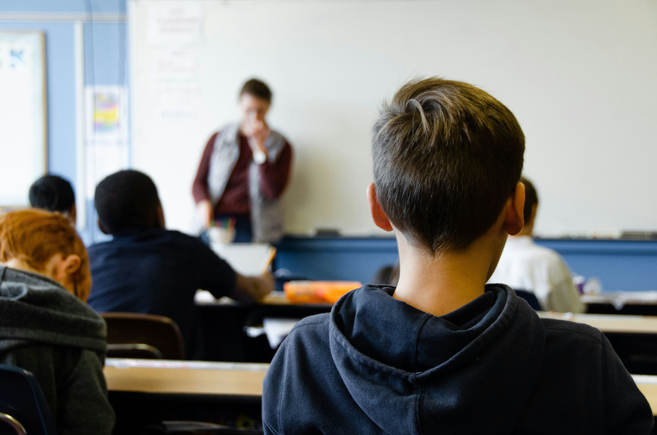 Jongetje met blauwe trui aan zit in klaslokaal. Voor hem zitten kinderen aan tafels en staat de leerkracht voor een whiteboard
